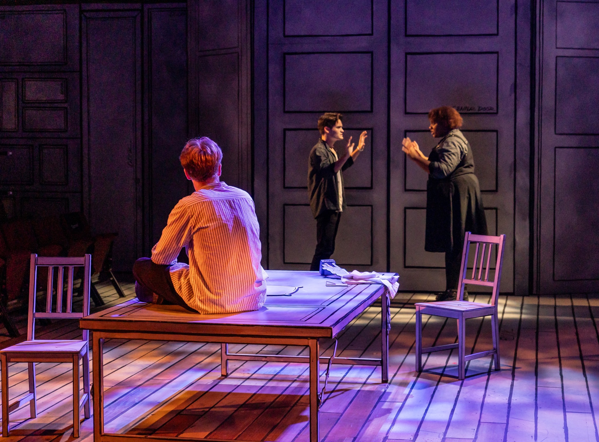 A seated performer faces two figures beneath scenic doors.
