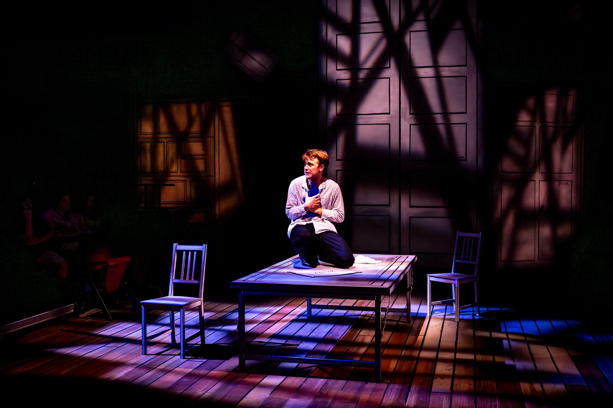 A performer kneels on a table amid patterned shadows.
