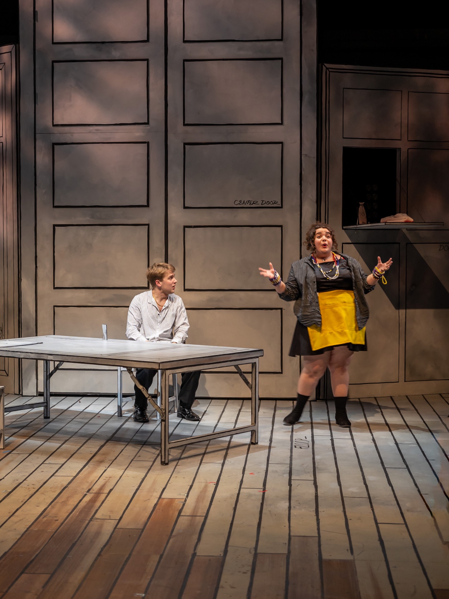 Two performers share a scene beside a white table and scenic doors.